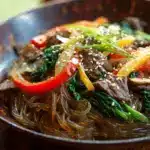 Colorful japchae bowl with glass noodles and sesame seeds on wood.