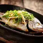 Whole Steamed Pompano with Ginger Sauce on a dark platter with scallions and ginger
