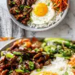 Overhead view of a vibrant Korean bulgogi rice bowl featuring glossy caramelized beef strips, a sunny-side-up egg with a runny yolk, fresh chopped scallions, shredded carrots, and a side of kimchi on a bed of fluffy white rice in a speckled ceramic bowl.