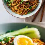 A close-up, top-down view of a vibrant bowl of chili crisp noodles. Glossy noodles are coated in red chili oil and flecked with ground pork. Bright green bok choy leaves and a halved soft-boiled egg sit on top, finished with a sprinkle of white sesame seeds and sliced green scallions.