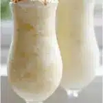 A tall hurricane glass filled with a thick, creamy frozen pina colada, garnished with a fresh pineapple wedge, a bright red maraschino cherry, and a sprinkle of toasted coconut flakes on a sunny outdoor table.