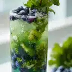 A backlit highball glass filled with a colorful layered kiwi blueberry mojito, featuring vibrant green kiwi slices and deep purple muddled blueberries, garnished with a fresh mint sprig and whole fruit on a dark, moody background.