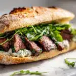 A ciabatta steak sandwich with creamy steak sandwich horseradish sauce and arugula.